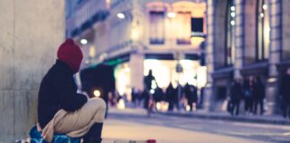 Une personne sans abri regardant vers des magasins est assise dans un boulevard haussmannien.